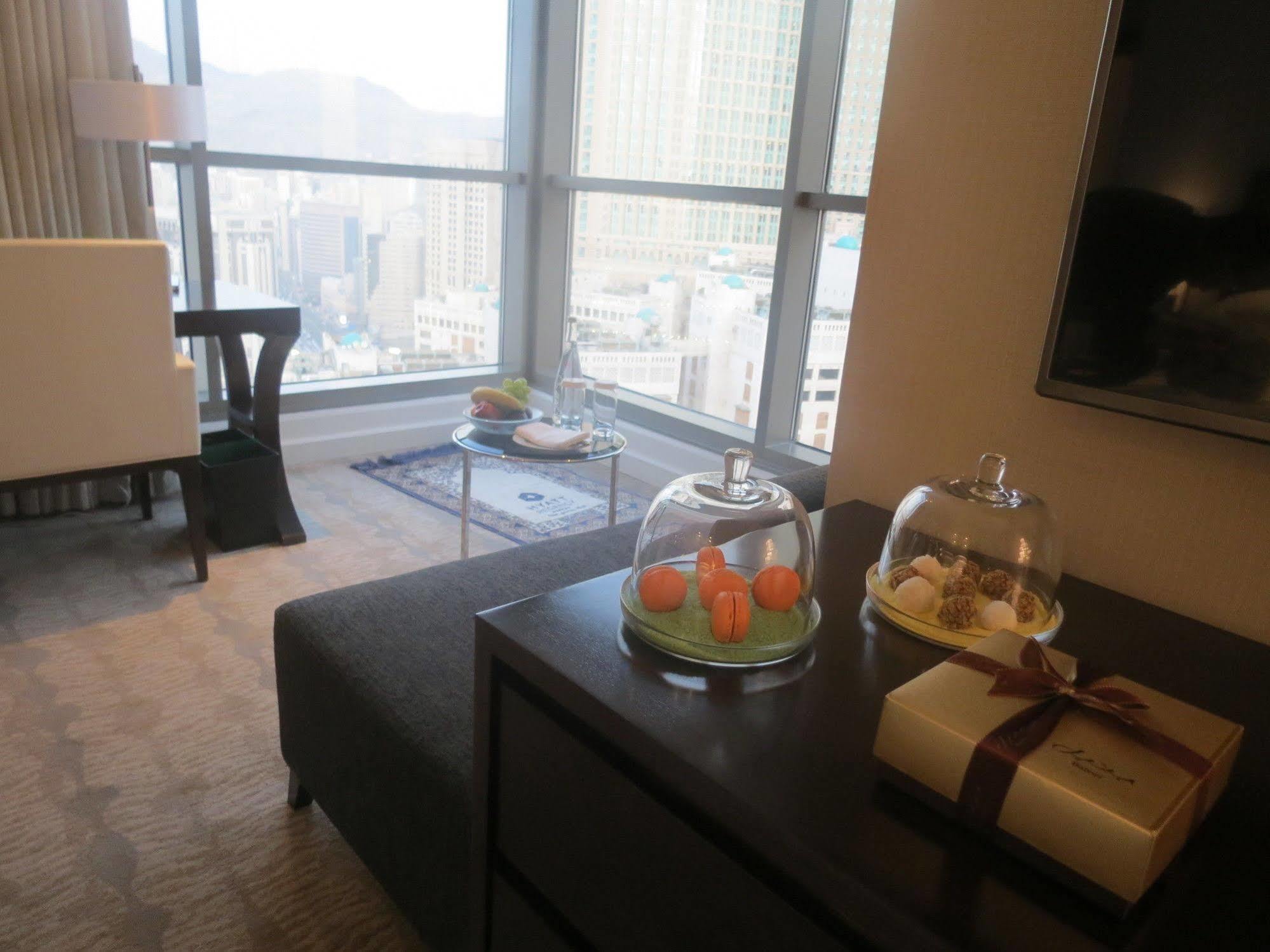 Hotel Jabal Omar Hyatt Regency Makkah Mekka Zewnętrze zdjęcie The photo shows a modern hotel room or a stylish living space with large windows offering a view of the city. In the foreground, there is a dark sofa and a low table with covered trays displaying what appears to be small desserts or delicacies. To th