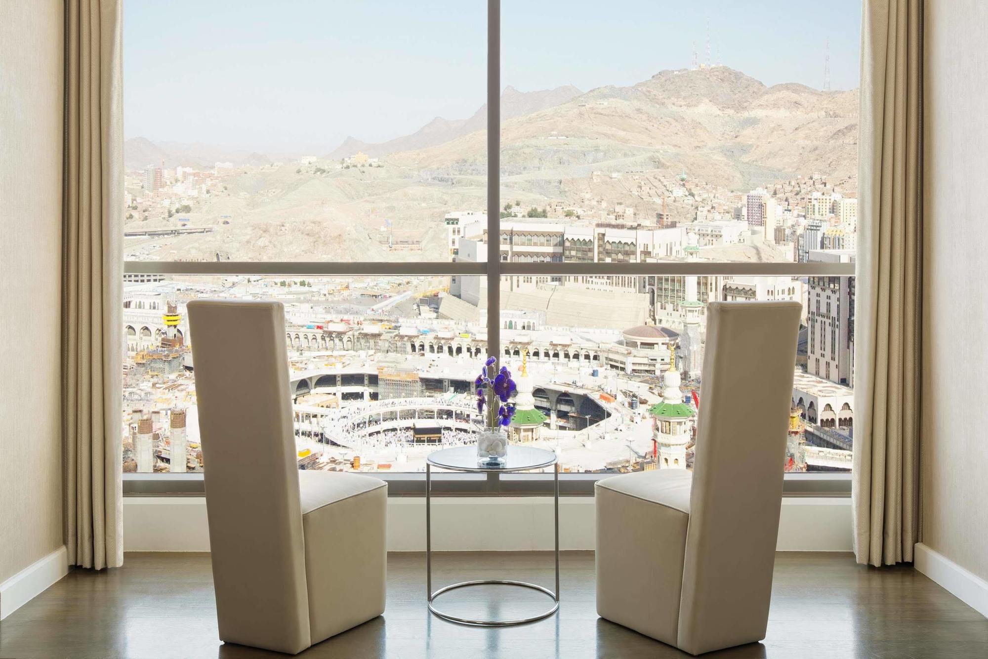 Hotel Jabal Omar Hyatt Regency Makkah Mekka Zewnętrze zdjęcie The photo shows a modern indoor setting with two tall, white chairs positioned facing a large window. Through the window, there is a panoramic view of a cityscape that includes mountainous terrain and buildings, suggesting it may be a notable locatio