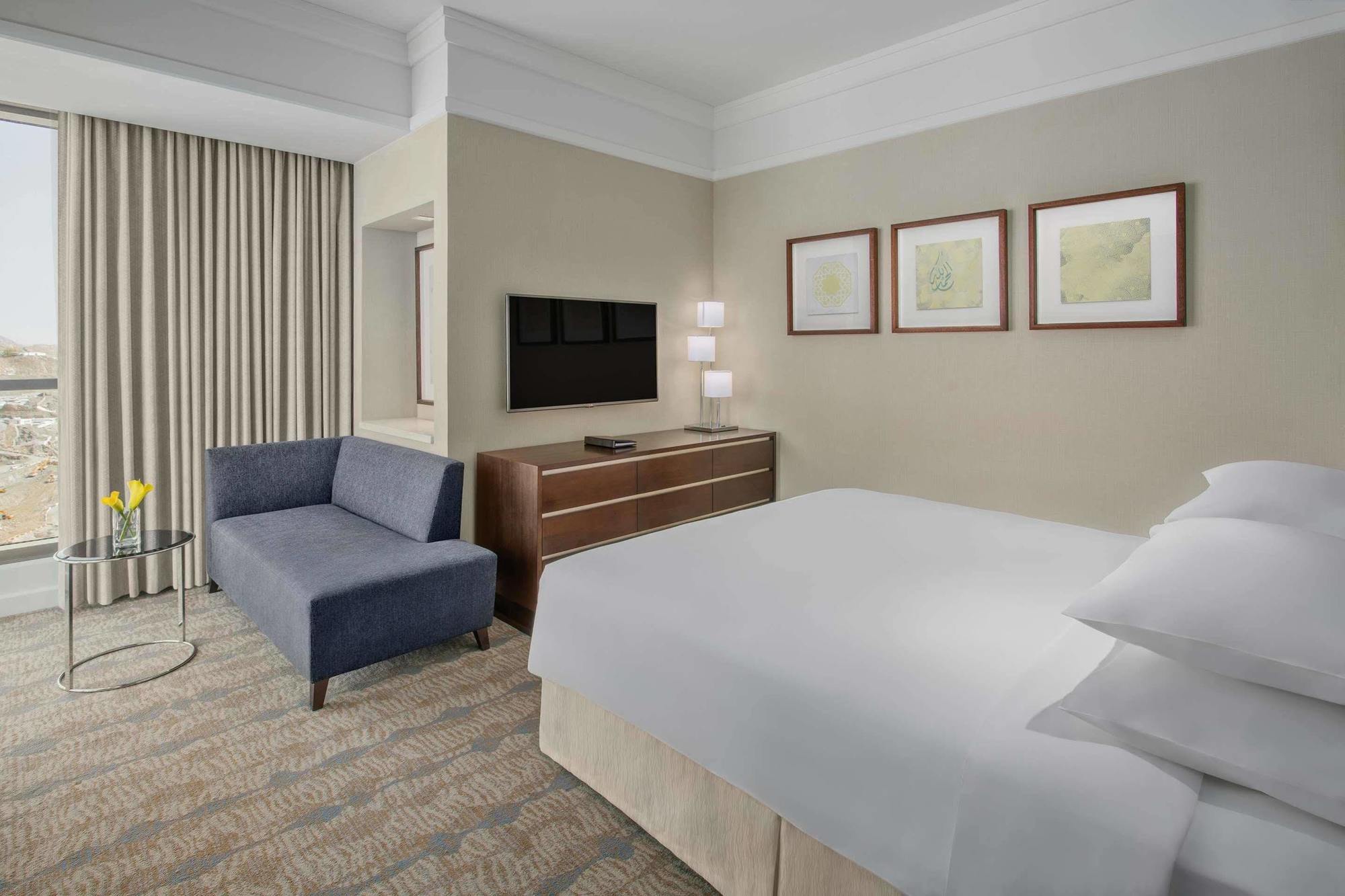 Hotel Jabal Omar Hyatt Regency Makkah Mekka Zewnętrze zdjęcie The photo shows a modern hotel room featuring a neatly made bed with white linens, a small blue couch to the side, and a wooden dresser with a television mounted on the wall above it. There are two framed pieces of art on the wall near the bed, and t